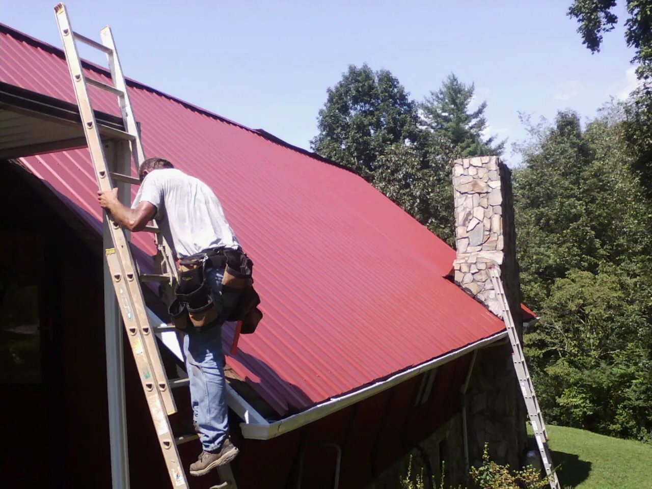 Roofer on a ladder beside a red metal roof and stone chimney — Soffit & Fascia Repair by Roofing Brothers in Faulkner